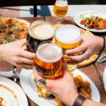 Vier mensen proosten met speciaalbieren aan een zomerse eettafel vol met pizza, friet, burgers en kip.