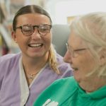 Zorgverlener en oudere vrouw lachen samen tijdens een huisbezoek, zorg aan huis als ondersteuning