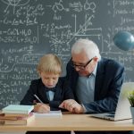 Mentorschap in actie: oudere man begeleidt jong kind bij wiskundeopdracht voor schoolbord vol formules.