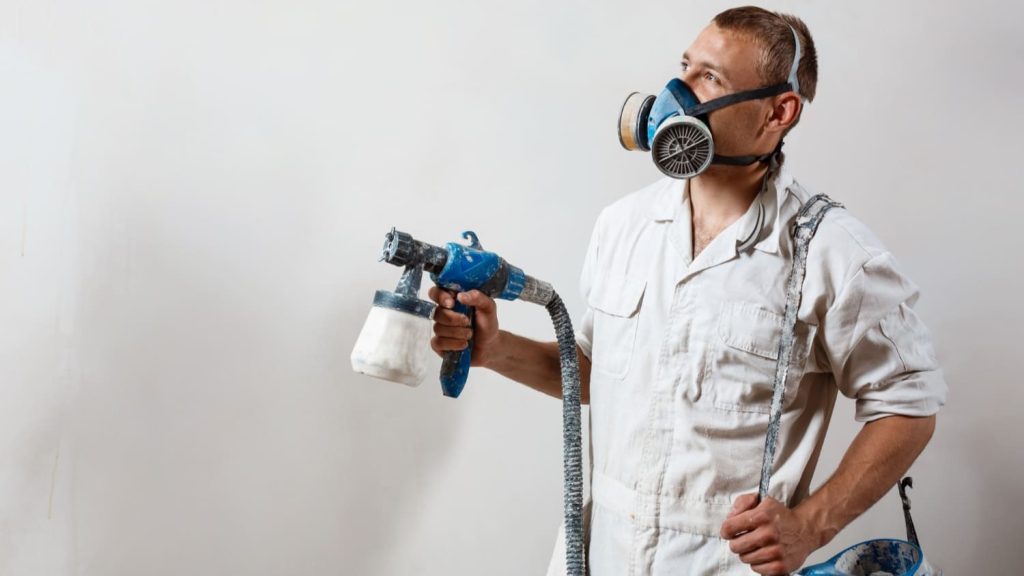 Schilder met spuitapparaat en beschermend masker voert periodiek onderhoud uit op muren.
